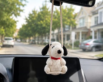 May include: A crocheted dog ornament with cream-colored yarn, black ears, a black nose, and a red collar. The figure is suspended by a light-colored cord from a rearview mirror, featuring a seated posture with visible yarn stitches throughout the body.