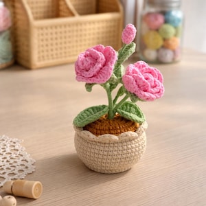 May include: A handmade crochet flower arrangement featuring two pink roses, green leaves, and a bud in a beige pot. The pot has a scalloped edge and sits on a wooden surface. The flowers are a vibrant pink.