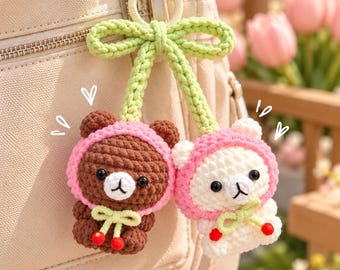 May include: A crochet keychain featuring two small bear figures, one brown and one white, each wearing a pink hood. The bears are connected by a light green crocheted stem with a bow at the top and small red berry accents at the base.
