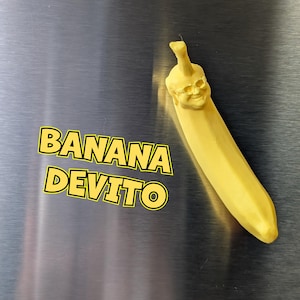 May include: A yellow banana with a face sculpted on the top, wearing sunglasses. The banana is on a silver surface with the text "BANANA DEVITO" in yellow letters.