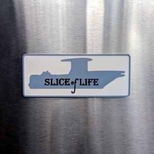 May include: A rectangular magnet with a white background and a blue boat silhouette. The text "SLICE of LIFE" is in black, with the "f" in "of" extending as a decorative flourish. The magnet is on a metallic surface.