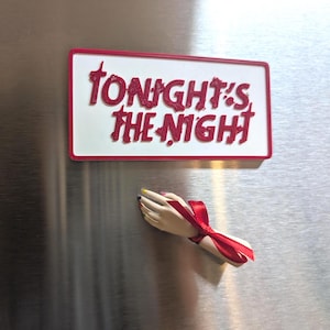 May include: A white rectangular magnet with a red border and the text "TONIGHT'S THE NIGHT" in red. Below the magnet is a small, decorative hand with a red ribbon tied around the wrist and painted nails.