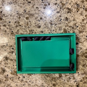 May include: A rectangular, bright green tray with a textured surface. The tray has a raised border and contains several small, black cylindrical objects. The tray is resting on a speckled, brown surface.