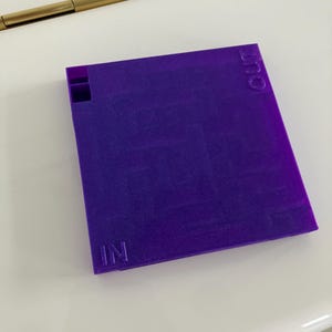 May include: A square, purple object with the word "IN" and "OUT" embossed on opposite sides. The object has a small rectangular cutout on one corner. The object is sitting on a white surface.