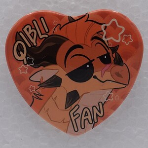 Puede incluir: Un pin en forma de coraz&oacute;n con una ilustraci&oacute;n de estilo dibujo de un drag&oacute;n color canela con cuernos oscuros y expresi&oacute;n satisfecha. El fondo es naranja con un patr&oacute;n sutil y estrellas blancas. El texto QIBLI FAN est&aacute; en letras negras.