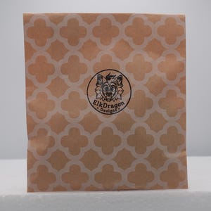 Puede incluir: Una bolsa de papel cuadrada de color marr&oacute;n con un patr&oacute;n repetitivo de cuatrifolios blancos. Centrado en la bolsa hay un sello de tinta negra circular que muestra una cara de animal estilizada con orejas grandes y el texto ElkDragon Designs.
