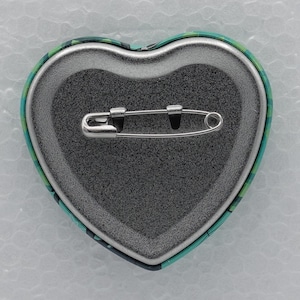 Puede incluir: Un bot&oacute;n de solapa en forma de coraz&oacute;n con un mecanismo de imperdible de metal plateado fijado a una base texturizada de color gris oscuro. El borde exterior del bot&oacute;n muestra un patr&oacute;n de cuadros verdes y negros visible en el contorno.