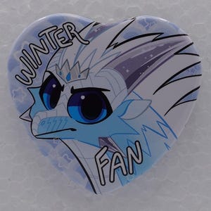 Puede incluir: Un pin en forma de coraz&oacute;n con una ilustraci&oacute;n estilizada de un drag&oacute;n azul claro y blanco con grandes ojos azul oscuro. El drag&oacute;n lleva una peque&ntilde;a corona y el texto WINTER FAN est&aacute; impreso en letras negras en los bordes superior e inferior.