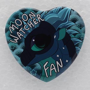 Puede incluir: Un pin en forma de coraz&oacute;n con un drag&oacute;n azul oscuro de ojos grandes. El fondo es un patr&oacute;n azul y turquesa de olas y peque&ntilde;as estrellas. El texto MOON WATCHER FAN est&aacute; impreso en letras blancas en la parte superior e inferior del dise&ntilde;o.
