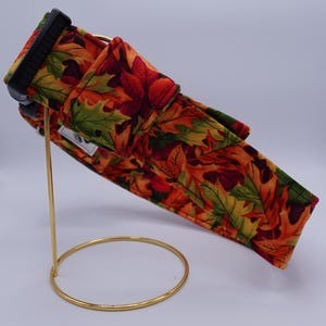 Könnte beinhalten: Ein Haustierhalsband aus Stoff mit einem herbstlichen Blättermuster in Orange, Rot, Gelb und Grün. Das Halsband verfügt über eine schwarze Kunststoff-Steckschnalle, einen Metallring und ein kleines, weißes, rechteckiges Markenetikett an der Seite.