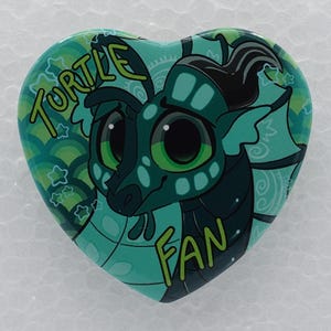Puede incluir: Un pin en forma de coraz&oacute;n con una criatura marina de dibujos animados con grandes ojos verdes y escamas de color verde azulado oscuro. El fondo muestra un patr&oacute;n de escamas verde claro y estrellas. El texto TURTLE FAN est&aacute; en letras amarillas.