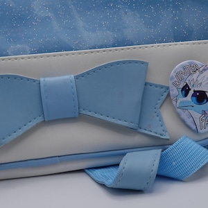 Puede incluir: Una mochila de color azul claro y blanco con un gran lazo azul claro en el panel frontal. Un pin en forma de coraz&oacute;n est&aacute; sujeto al lateral, mostrando un personaje de drag&oacute;n de dibujos animados con el texto "WINTER FAN" en letra estilizada.