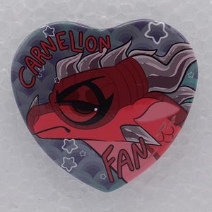 Puede incluir: Un pin en forma de coraz&oacute;n con un personaje de drag&oacute;n rojo con cuernos grises y una expresi&oacute;n severa. El fondo es un patr&oacute;n azul oscuro con peque&ntilde;as estrellas blancas. El texto CARNELION FAN est&aacute; impreso en letras rojas alrededor del drag&oacute;n.