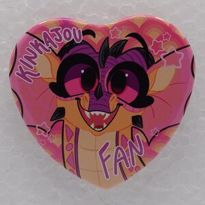 Puede incluir: Un pin en forma de coraz&oacute;n con una ilustraci&oacute;n de dibujos animados de una criatura amarilla y morada con ojos grandes y una amplia sonrisa. El fondo es un degradado rosa con arcos de arco&iacute;ris y estrellas. El texto KINKAJOU FAN est&aacute; en blanco.