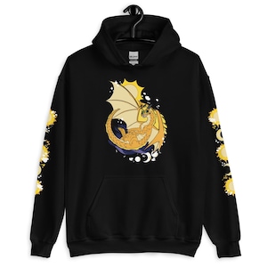 Könnte beinhalten: Schwarzer Kapuzenpullover mit einem Cartoon-Drachendesign in Gelb- und Orangetönen, mit gelben Sonnen- und Mondakzenten. Die Ärmel haben ein sich wiederholendes Sonnendesign. Der Pullover hat eine Vordertasche und eine Kapuze mit Kordelzug.