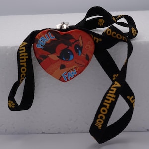 Puede incluir: Una insignia en forma de coraz&oacute;n con una ilustraci&oacute;n de un drag&oacute;n rojo y naranja con el texto azul PERIL FAN, unida a un cord&oacute;n negro con huellas amarillas y el texto Anthrocon impreso en amarillo repetidamente en la cinta de tela.