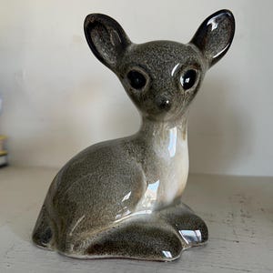May include: A ceramic figurine of a deer in a seated position. The deer has large, dark eyes, and its fur is a mix of gray and brown with glossy highlights. The ears are dark brown, and the figurine has a smooth, glazed finish.