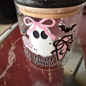 Puede incluir: Un tarro de cristal transparente con una tapa negra decorada con rosas rosas y un diseño de fantasma blanco y rosa. El tarro tiene un borde de madera y presenta las palabras "demure spooky". También hay un murciélago y un ataúd.