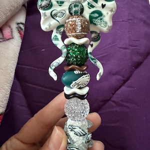 May include: A decorative pen featuring a white bow topper with Philadelphia Eagles logos, football helmets, and stars. The barrel is adorned with a series of beads including a brown football, a green rhinestone bead, and white eagle logo beads.