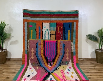 May include: A vibrant, hand-woven rug with a geometric design. The rug features a mix of colors, including pink, orange, blue, and green. The design includes abstract shapes and patterns, with a fringed edge. The rug is displayed on a wooden floor.