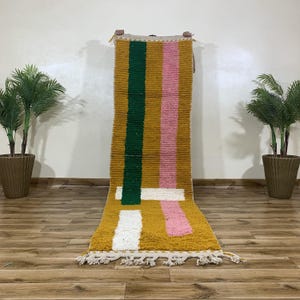 May include: Mustard yellow and multi-colored rug with green and pink vertical stripes and white horizontal accents. The rug has a textured, woven appearance and a fringed edge. The rug is displayed on a wooden floor.