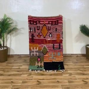 May include: A rectangular, hand-woven rug with a vibrant, geometric design. The rug features a mix of colors, including red, orange, green, and black, with a variety of shapes and patterns. The rug is held up against a white wall, showcasing its intricate details.