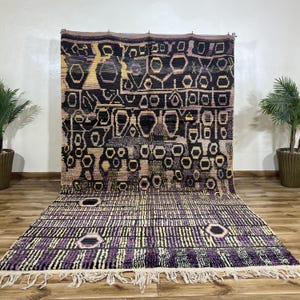 Puede incluir: Una alfombra rectangular grande con un patrón geométrico en negro, morado y amarillo. La alfombra tiene flecos en el borde inferior y se muestra contra una pared neutra. El diseño incluye varias formas y líneas.