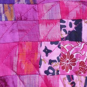 May include: Close-up of a patchwork textile with a quilted design. The fabric features a variety of pink and purple hues, with floral and geometric patterns. The stitching creates a grid-like pattern across the surface.
