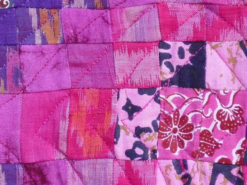 May include: Close-up of a patchwork textile with a quilted design. The fabric features a variety of pink and purple hues, with floral and geometric patterns. The stitching creates a grid-like pattern across the surface.