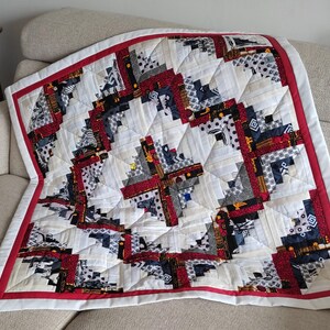 May include: A handmade quilt with a log cabin design. The quilt features a white background with red, black, and gray patterned squares. A red border frames the quilt, which is folded on a beige sofa. The quilt is a cozy home decor item.