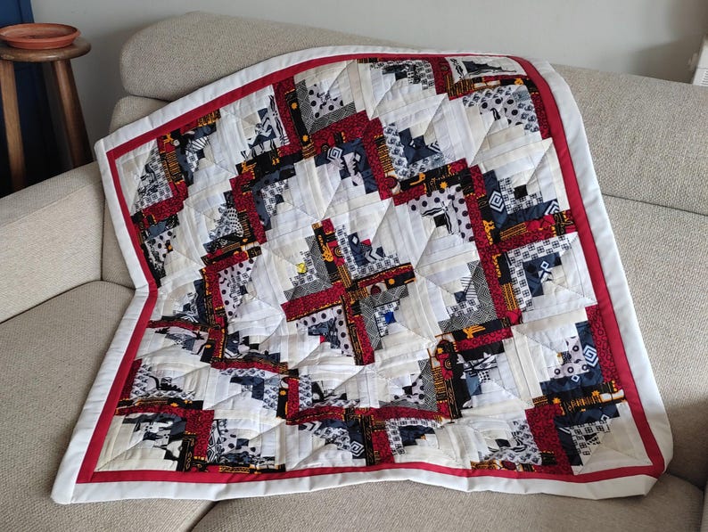 May include: A handmade quilt with a log cabin design. The quilt features a white background with red, black, and gray patterned squares. A red border frames the quilt, which is folded on a beige sofa. The quilt is a cozy home decor item.
