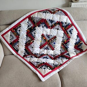May include: A handmade quilt with a geometric pattern. The quilt features a white background with red, navy, and gray accents. A red border frames the quilt, which is placed on a beige sofa. The quilt is square-shaped and appears to be made of fabric scraps.