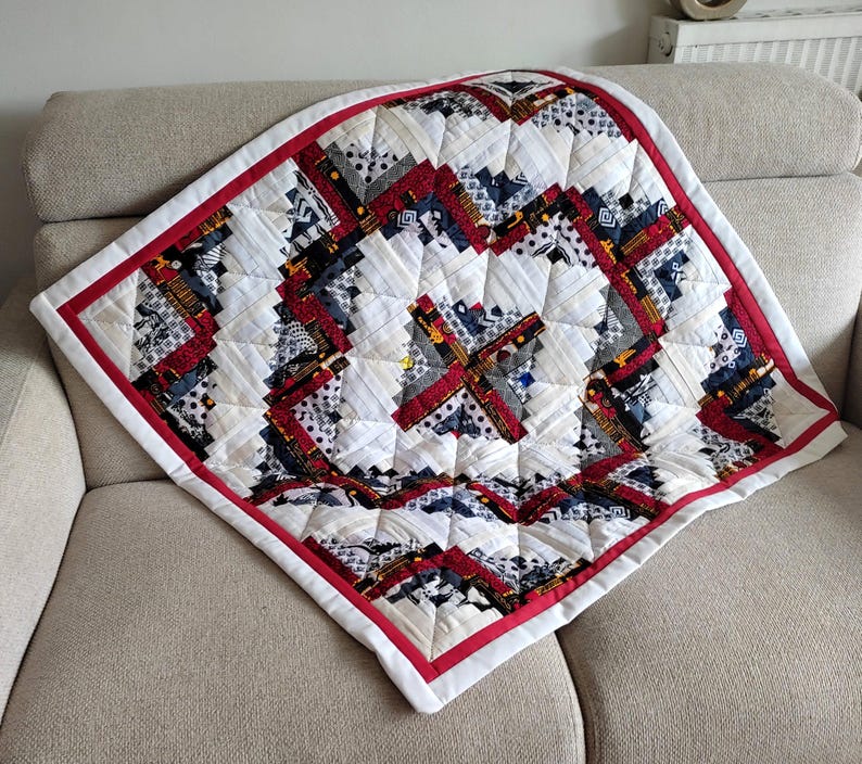 May include: A handmade quilt with a geometric pattern. The quilt features a white background with red, navy, and gray accents. A red border frames the quilt, which is placed on a beige sofa. The quilt is square-shaped and appears to be made of fabric scraps.