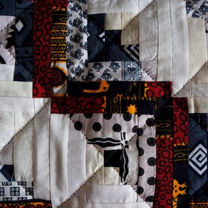 May include: Close-up of a handmade quilt with a geometric pattern. The quilt features squares and rectangles in white, black, red, and patterned fabrics. The design includes repeating shapes and stitched lines.