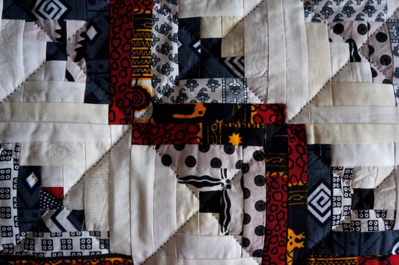May include: Close-up of a handmade quilt with a geometric pattern. The quilt features squares and rectangles in white, black, red, and patterned fabrics. The design includes repeating shapes and stitched lines.