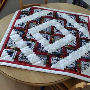 May include: A square, handmade quilt with a log cabin design. The quilt features a white background with blocks of red, navy, and patterned fabrics. A red border frames the quilt, which is placed on a round wooden table.