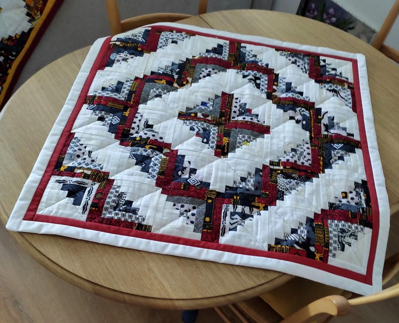 May include: A square, handmade quilt with a log cabin design. The quilt features a white background with blocks of red, navy, and patterned fabrics. A red border frames the quilt, which is placed on a round wooden table.