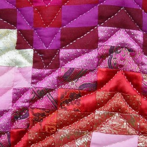 May include: Close-up of a patchwork quilt with a geometric design. The quilt features squares and triangles in shades of pink, red, and purple. The fabric has a satin sheen and is stitched with white thread. Some sections have gold accents.