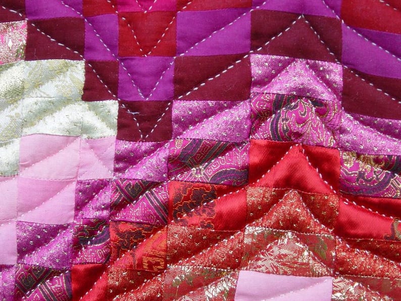 May include: Close-up of a patchwork quilt with a geometric design. The quilt features squares and triangles in shades of pink, red, and purple. The fabric has a satin sheen and is stitched with white thread. Some sections have gold accents.