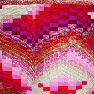 May include: A close-up of a handmade quilt with a vibrant colour palette. The quilt features a patchwork design with squares and geometric patterns in shades of red, pink, and gold. The quilt's texture and intricate stitching are visible.