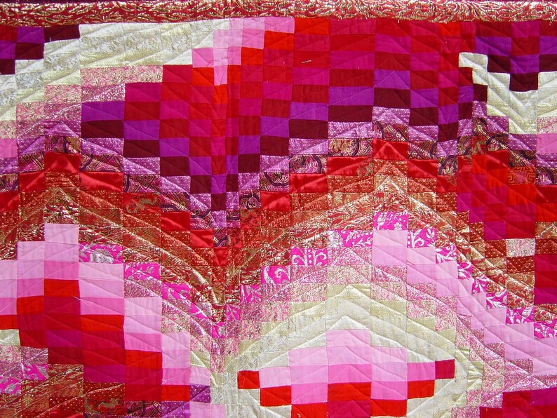 May include: A close-up of a handmade quilt with a vibrant colour palette. The quilt features a patchwork design with squares and geometric patterns in shades of red, pink, and gold. The quilt's texture and intricate stitching are visible.