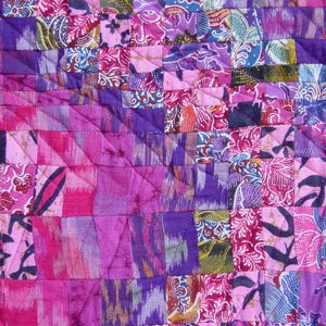 May include: Close-up of a patchwork quilt with a vibrant mix of pink, purple, and blue fabrics. The quilt features a geometric pattern of squares and rectangles, with floral and abstract designs. The stitching is visible, adding texture.
