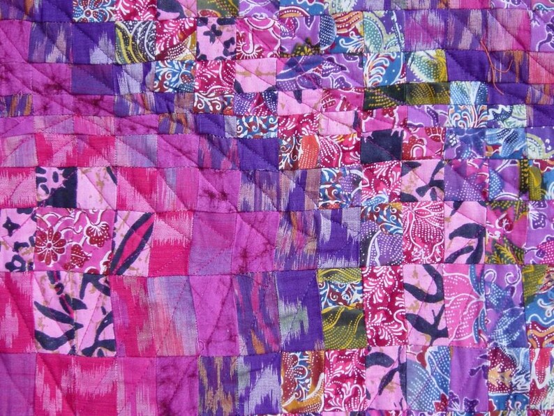 May include: Close-up of a patchwork quilt with a vibrant mix of pink, purple, and blue fabrics. The quilt features a geometric pattern of squares and rectangles, with floral and abstract designs. The stitching is visible, adding texture.