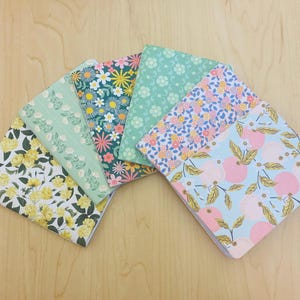 May include: A collection of notebooks with floral patterns. The notebooks feature various designs, including yellow roses, daisies, and pink citrus fruits. The covers are in shades of green, pink, and blue.