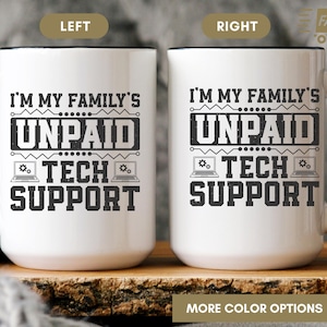 May include: White ceramic mug with a black handle and rim. The mug features the text "I'M MY FAMILY'S UNPAID TECH SUPPORT" in a bold, black font. The design includes laptop icons. The mug is on a wooden surface.