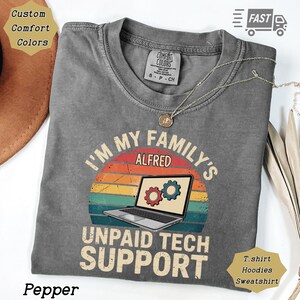 May include: A gray t-shirt with the text "I'M MY FAMILY'S ALFRED UNPAID TECH SUPPORT" and a laptop graphic with gears. The shirt has a vintage sunset design and the word "Pepper" at the bottom. A necklace is draped across the shirt.