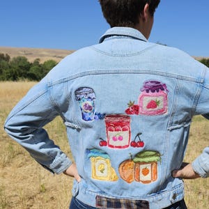Può includere: Giacca di jeans azzurra con disegni di barattoli di marmellata ricamati sul retro. La giacca presenta varie conserve di frutta, tra cui fragole e ciliegie. La giacca è indossata all'aperto in una giornata di sole.