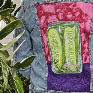 Pode incluir: Casaco de ganga azul claro com um desenho em feltro de um frasco de picles nas costas. O frasco de picles é verde com detalhes amarelos, sobre um fundo rosa e roxo. O casaco tem gola clássica e fecho de botões.