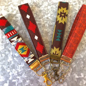 May include: Five colorful keychains with different geometric patterns. Each keychain has a gold metal ring and a silver metal clasp. The keychains are on a silver background.