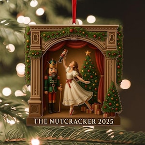 Könnte beinhalten: Weihnachtsschmuck mit einer Szene aus dem Ballett "Der Nussknacker". Es zeigt eine Ballerina, einen Nussknackersoldaten und einen Weihnachtsbaum. Der Text "THE NUTCRACKER 2025" steht unten. Der Schmuck ist mit rotem Band verziert.